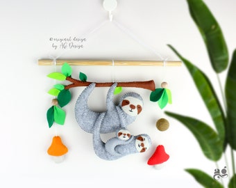 May include: A gray felt sloth with a baby sloth hanging on its back. The sloth is hanging from a wooden branch with green felt leaves. There are also two felt mushrooms, one orange and one red, hanging from the branch.