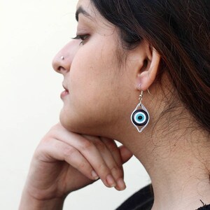 May include: Silver earrings with a blue and black evil eye charm. The earrings are dangling and have a silver hook closure.