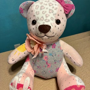 May include: A handmade patchwork teddy bear with a pink bow, floral accents, and a heart-shaped pattern on its head. The bear has a brown nose, black eyes, and a pink flower embellishment. The bear's body is made of various fabrics, including pink, gray, and white.