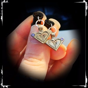 May include: A pair of silver heart-shaped earrings with a small purple gemstone on each. The earrings are hanging from black plugs.