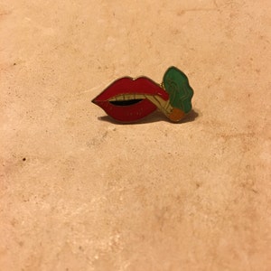 May include: A red enamel pin in the shape of a mouth with a gold outline. The mouth is open, revealing teeth, and is holding a green and orange object. The pin is on a light brown surface.