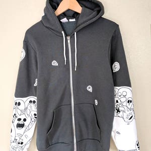 May include: A dark gray zip-up hoodie with white cartoon ghost and skull designs on the sleeves. The hoodie has a white zipper and a white drawstring.