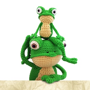 May include: Two crocheted green frogs, one sitting on top of the other. The larger frog has large, round eyes and is sitting with its arms crossed. The smaller frog is sitting on the larger frog's head with its arms outstretched.