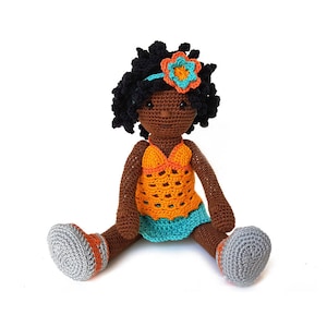 Op de afbeelding: Een gehaakte pop met bruine huid, zwart krullend haar en een blauwe en oranje jurk. De pop draagt ​​een bloemenhoofdband en grijze schoenen.