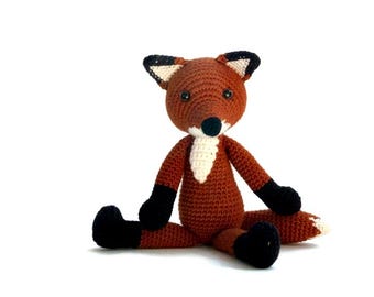 May include: A crocheted fox plush toy with brown fur, black ears, a white chest, and black feet. The fox is sitting with its legs spread out.