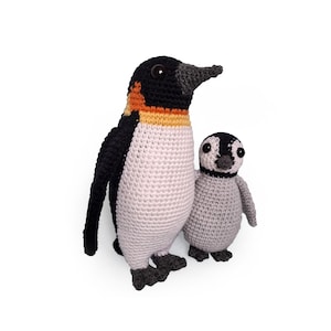 Op de afbeelding: Twee gehaakte pinguïnfiguren. De grotere pinguïn heeft een zwarte kop, vleugels en voeten, met een witte buik en oranje en gele accenten. De kleinere pinguïn is grijs met zwarte markeringen. Beide hebben grote, donkere ogen.