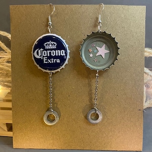 Puede incluir: Pendientes hechos a mano con tapones de botella Corona Extra reciclados. Uno muestra el logotipo de Corona Extra sobre un fondo azul marino. El otro tiene una estrella rosa y pequeños detalles de estrellas. Ambos cuelgan de cadenas plateadas.