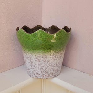 Op de afbeelding: Een keramische bloempot met een geschulpte rand. De bovenkant is groen, overgaand in een gespikkelde wit en bruine basis. De rand is donkerbruin. De pot staat op een witte plank tegen een roze muur.