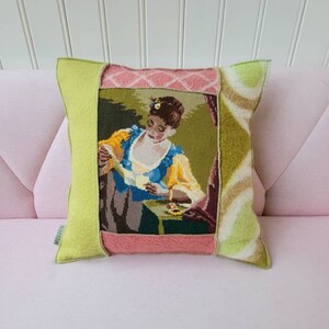 Op de afbeelding: Een groen, roze en bruin patchwork kussen met een naaldkunst afbeelding van een vrouw in een blauwe jurk die een brief leest. Het kussen heeft een groen label met de tekst "HEBBIEN".