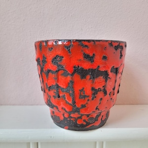 May include: A red and black ceramic plant pot with a textured, lava-like design. The pot has a cylindrical shape with a slightly flared rim and a dark base. The exterior features a vibrant red glaze with black accents, creating a striking contrast.