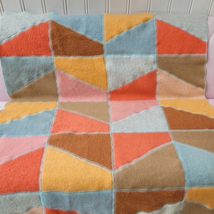 Puede incluir: Una manta de patchwork con una disposición geométrica de paneles de tela afieltrada en tonos naranja, amarillo, marrón, azul claro y rosa suave. Las secciones individuales están unidas con costuras decorativas visibles en azul claro.