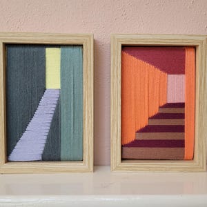 Op de afbeelding: Twee rechthoekige wandkunstwerken met geometrische draadkunstontwerpen in lichte houten lijsten. Het linker stuk toont donkergrijs, lichtgroen, geel en lavendelkleurig garen. Het rechter stuk bevat oranje, roze en donkerbruin garen.