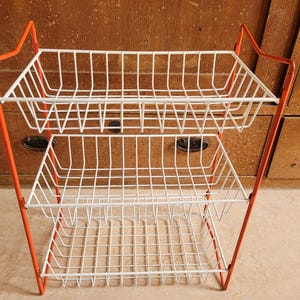 May include: A two-tiered wire basket stand with a bright orange frame and white wire baskets. The stand has a retro design, ideal for kitchen or bathroom storage. The baskets are rectangular and the stand is approximately 61 cm tall.