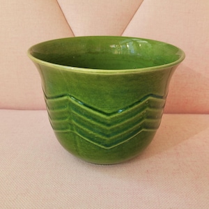 May include: A glossy, grass-green ceramic planter featuring a wide, flared rim and a series of horizontal, V-shaped chevron ridges embossed around the exterior body. The vessel has a smooth, reflective finish and a deep, open interior basin.