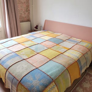 Könnte beinhalten: Eine Patchwork-Decke in Pastellfarben wie Gelb, Blau und Rosa bedeckt ein Bett. Die Decke besteht aus quadratischen Flicken, die mit orangefarbenen Nähten zusammengenäht sind. Das Bett hat ein rosa Kopfteil.