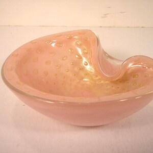 May include: A pink Murano glass bowl with a unique, asymmetrical shape. The bowl features a light pink hue with embedded gold-colored speckles. The rim curves gracefully, adding to its artistic design. A decorative piece for home decor.