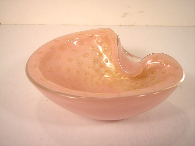May include: A pink Murano glass bowl with a unique, asymmetrical shape. The bowl features a light pink hue with embedded gold-colored speckles. The rim curves gracefully, adding to its artistic design. A decorative piece for home decor.
