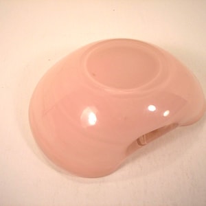 May include: A light pink, rounded glass bowl with a small, circular indentation in the center. The bowl has a smooth, glossy surface and a small, curved opening on one side.
