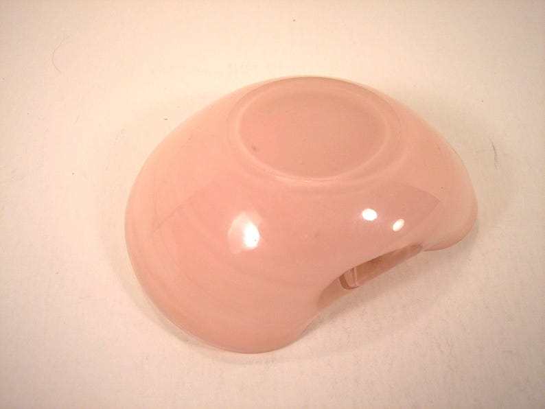 May include: A light pink, rounded glass bowl with a small, circular indentation in the center. The bowl has a smooth, glossy surface and a small, curved opening on one side.