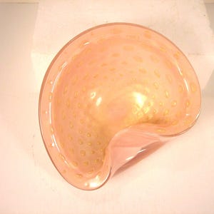 May include: A peach-colored, glass bowl with a unique, asymmetrical shape. The bowl features small, golden-yellow speckles throughout the glass. The rim of the bowl is smooth and the overall design is elegant and decorative.