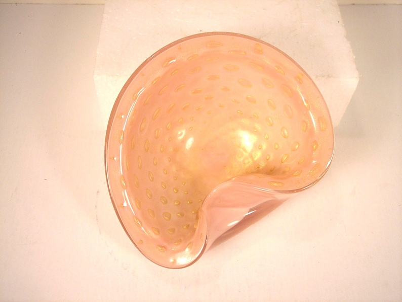 May include: A peach-colored, glass bowl with a unique, asymmetrical shape. The bowl features small, golden-yellow speckles throughout the glass. The rim of the bowl is smooth and the overall design is elegant and decorative.