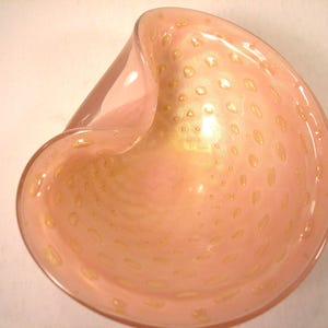 May include: A pink glass bowl with a unique, asymmetrical shape. The bowl has a speckled design with gold-colored accents. The interior of the bowl has a glossy finish, and the exterior is smooth.