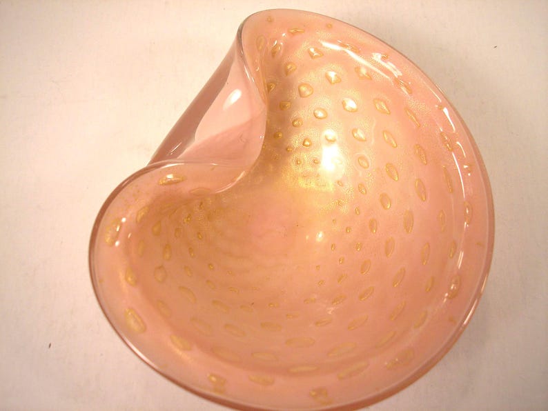 May include: A pink glass bowl with a unique, asymmetrical shape. The bowl has a speckled design with gold-colored accents. The interior of the bowl has a glossy finish, and the exterior is smooth.