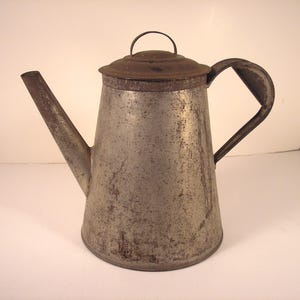 May include: Vintage metal coffee pot with a spout, handle, and lid. The pot has a silver finish with signs of wear and rust. The handle is curved, and the lid has a small loop. A classic kitchenware item.