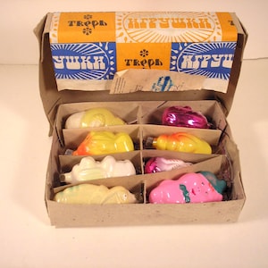 May include: A vintage box of Christmas ornaments. The box is brown with a colorful lid featuring text in a foreign language. Inside, six compartments hold colorful, uniquely shaped ornaments in shades of yellow, pink, and purple.