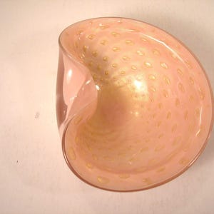 May include: A pink glass bowl with a unique, curved shape. The bowl has a speckled pattern of small, golden-colored dots throughout the interior. The glass has a glossy finish, reflecting light.