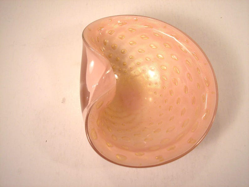 May include: A pink glass bowl with a unique, curved shape. The bowl has a speckled pattern of small, golden-colored dots throughout the interior. The glass has a glossy finish, reflecting light.