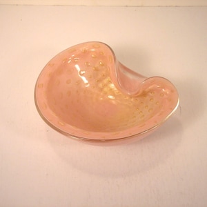 May include: A pink glass ashtray with a unique, curved shape. The ashtray has a light pink hue with gold-colored speckles throughout the glass. The rim of the ashtray is smooth and rounded.