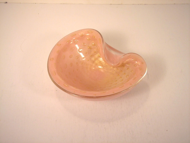 May include: A pink glass ashtray with a unique, curved shape. The ashtray has a light pink hue with gold-colored speckles throughout the glass. The rim of the ashtray is smooth and rounded.