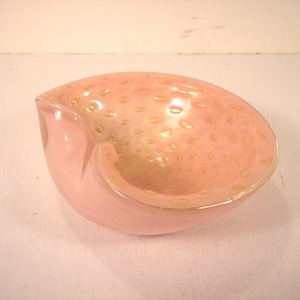 May include: A pink glass ashtray with a unique shape. The interior has a speckled design with gold-colored accents. The ashtray has a smooth, glossy finish and a curved, open design.