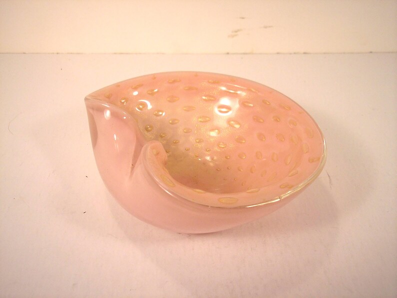 May include: A pink glass ashtray with a unique shape. The interior has a speckled design with gold-colored accents. The ashtray has a smooth, glossy finish and a curved, open design.