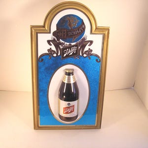 May include: Vintage Schlitz beer advertising sign. The sign features a framed design with a blue background and a bottle of Schlitz beer. The top of the sign has a globe with the word "Schlitz" on it. The frame is gold-colored.