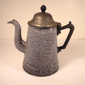 May include: Vintage gray and white enamelware coffee pot with a metal lid and handle. The pot has a spout and a decorative handle. The enamelware has a speckled pattern. The coffee pot is a kitchenware item.