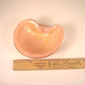 May include: A pink glass bowl with a unique shape and a textured surface. The bowl has a light pink color with small, golden-colored spots throughout. A wooden ruler is next to the bowl.