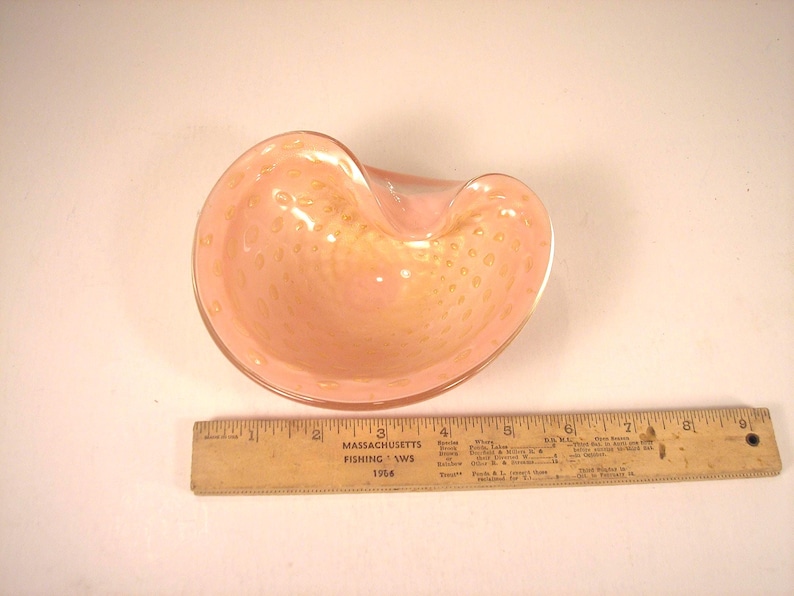 May include: A pink glass bowl with a unique shape and a textured surface. The bowl has a light pink color with small, golden-colored spots throughout. A wooden ruler is next to the bowl.