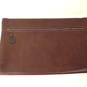 Op de afbeelding: Een bruine leren documententas met een ritssluiting en een kleine, donkere leren treklip. De tas heeft een rechthoekige vorm en een subtiel reliëflogo in de rechter benedenhoek. De ritssluiting is goudkleurig.