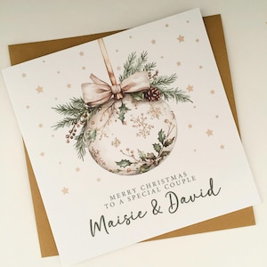 Può includere: Un biglietto di Natale con un'illustrazione ad acquerello di una palla di Natale, ornata da fiocchi di neve, agrifoglio e un fiocco. Il biglietto recita "Merry Christmas to a Special Couple" ed è indirizzato a Maisie & David. Il biglietto è su una busta marrone.