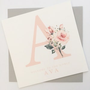 Può includere: Un biglietto rosa con una grande lettera A rosa decorata con un motivo floreale ad acquerello. Il testo "Welcome to the world AVA" è stampato sotto la lettera.
