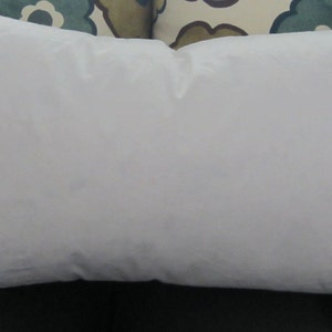 May include: A white pillow with a smooth, plain surface. The pillow is lying on a patterned fabric surface.
