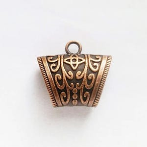 Op de afbeelding: Een koperkleurige metalen hanger met een versierd ontwerp. De hanger heeft een lus aan de bovenkant om aan een ketting of koord te bevestigen.