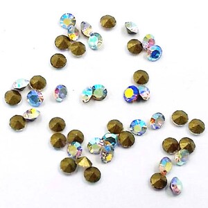 Op de afbeelding: Een assortiment iriserende en bronskleurige strass-steentjes verspreid op een witte ondergrond. De iriserende stenen reflecteren meerdere kleuren, terwijl de bronzen stenen een facetontwerp hebben. Ideaal voor knutselen en het maken van sieraden.