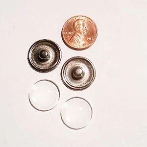 May include: An overhead shot shows two antique silver-colored snap fasteners, two clear, round, glass cabochons, and a copper penny. The penny is positioned near the fasteners, providing a size reference. The items are arranged on a white surface.