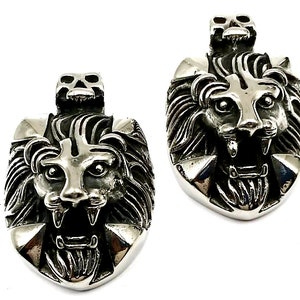 May include: Two silver lion head pendants with a skull detail on top. The lion heads are detailed with a cross on the forehead and a mane of hair.