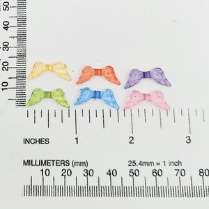 May include: Six pairs of colorful angel wings made of plastic. The wings are approximately 1 inch in length and are arranged in a row on a white surface with a ruler for scale.