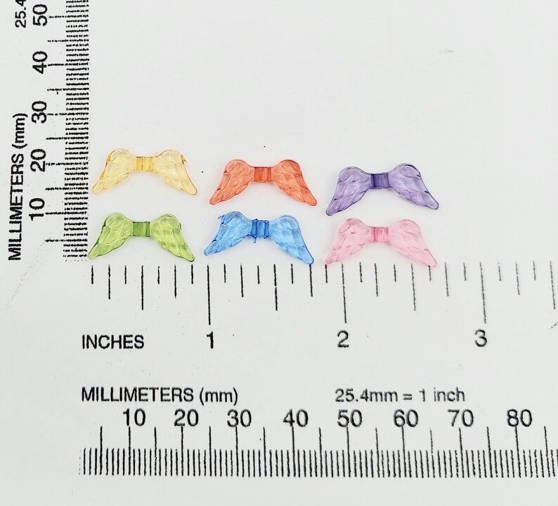 May include: Six pairs of colorful angel wings made of plastic. The wings are approximately 1 inch in length and are arranged in a row on a white surface with a ruler for scale.