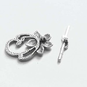 May include: Silver-toned metal clasp with a floral design. The clasp is made of two pieces, one with a decorative floral design and the other with a simple bar.